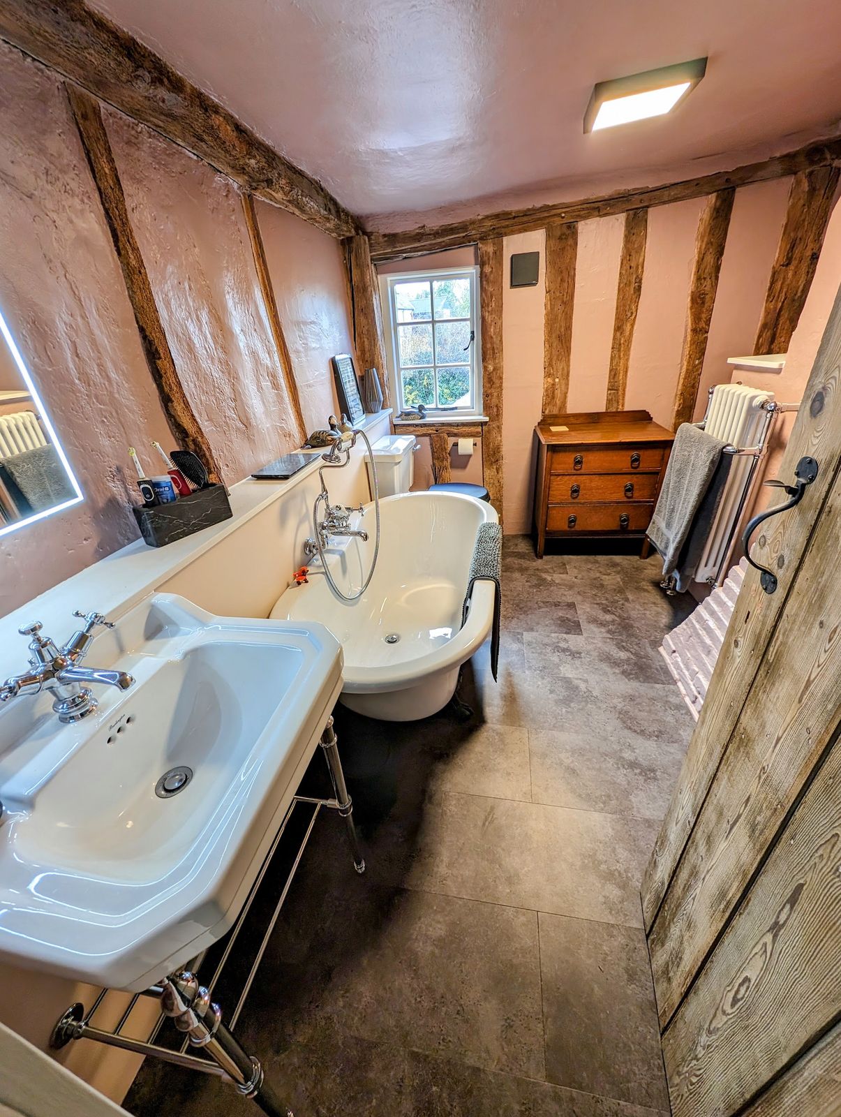 A bathroom under oak beams with twin basins, a roll-top bath and a column radiator