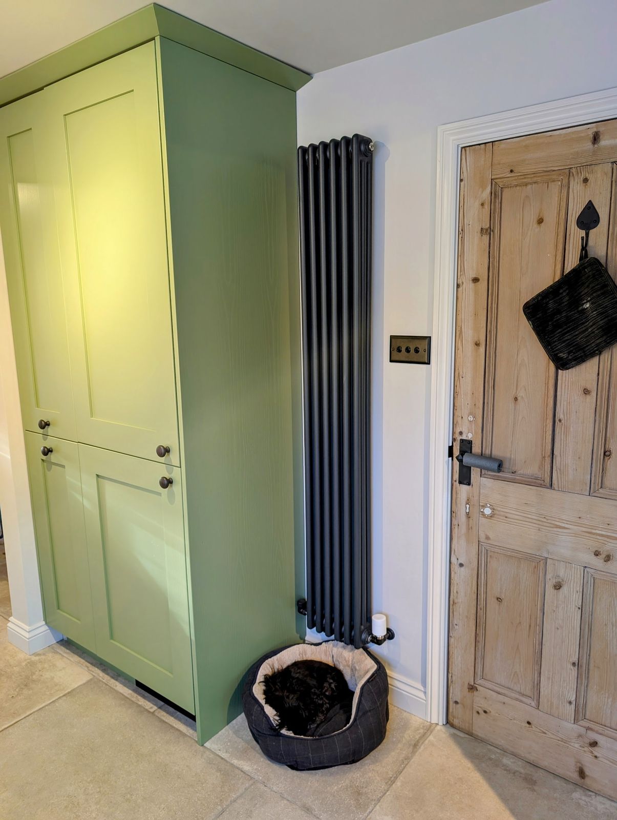 A tall anthracite column radiator beside a green cupboard and a cottage door